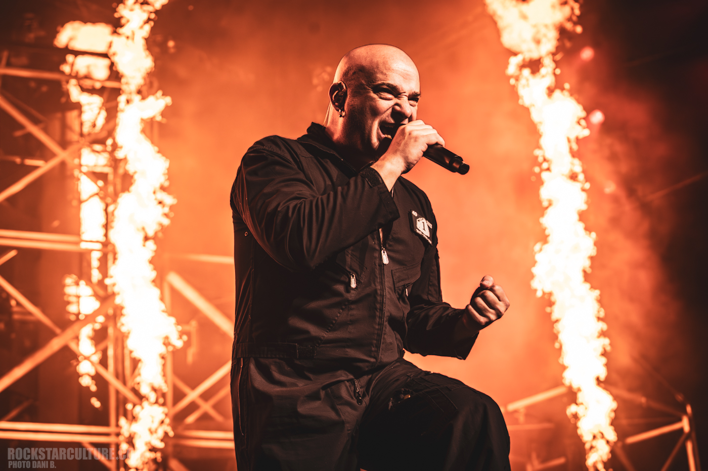 A singer performing passionately on stage with flames in the background, dressed in a black jumpsuit, captured during a live concert.
