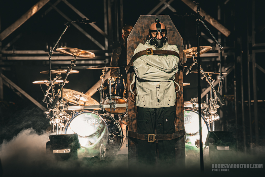 A performer dressed in a straitjacket stands inside a large coffin-like structure on stage, surrounded by drum sets and atmospheric smoke during a Disturbed concert.
