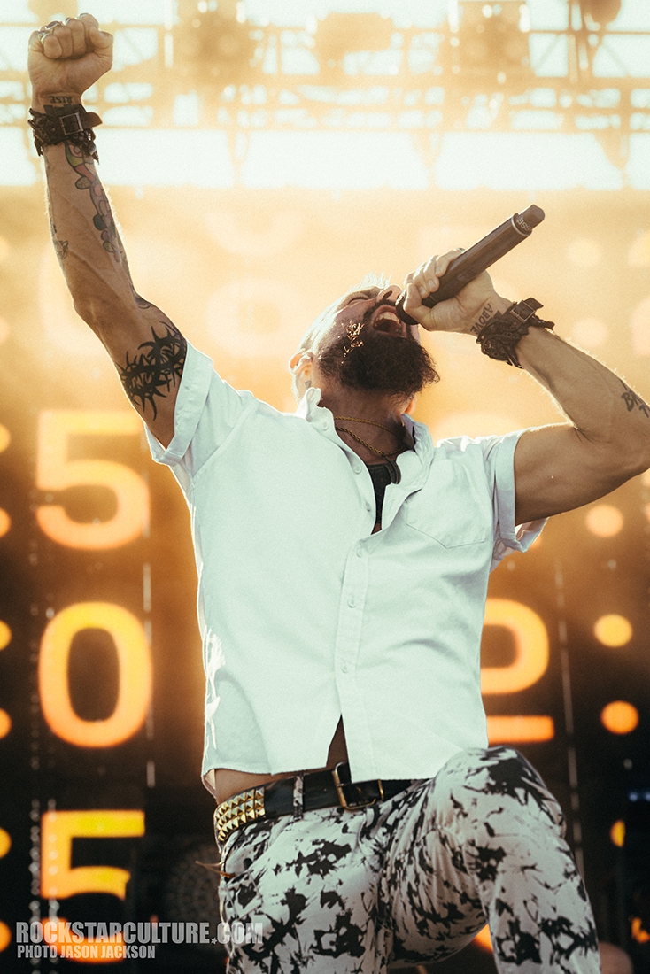 A male singer passionately performing on stage, holding a microphone and raising his arm, with bright stage lights and a large digital display in the background.