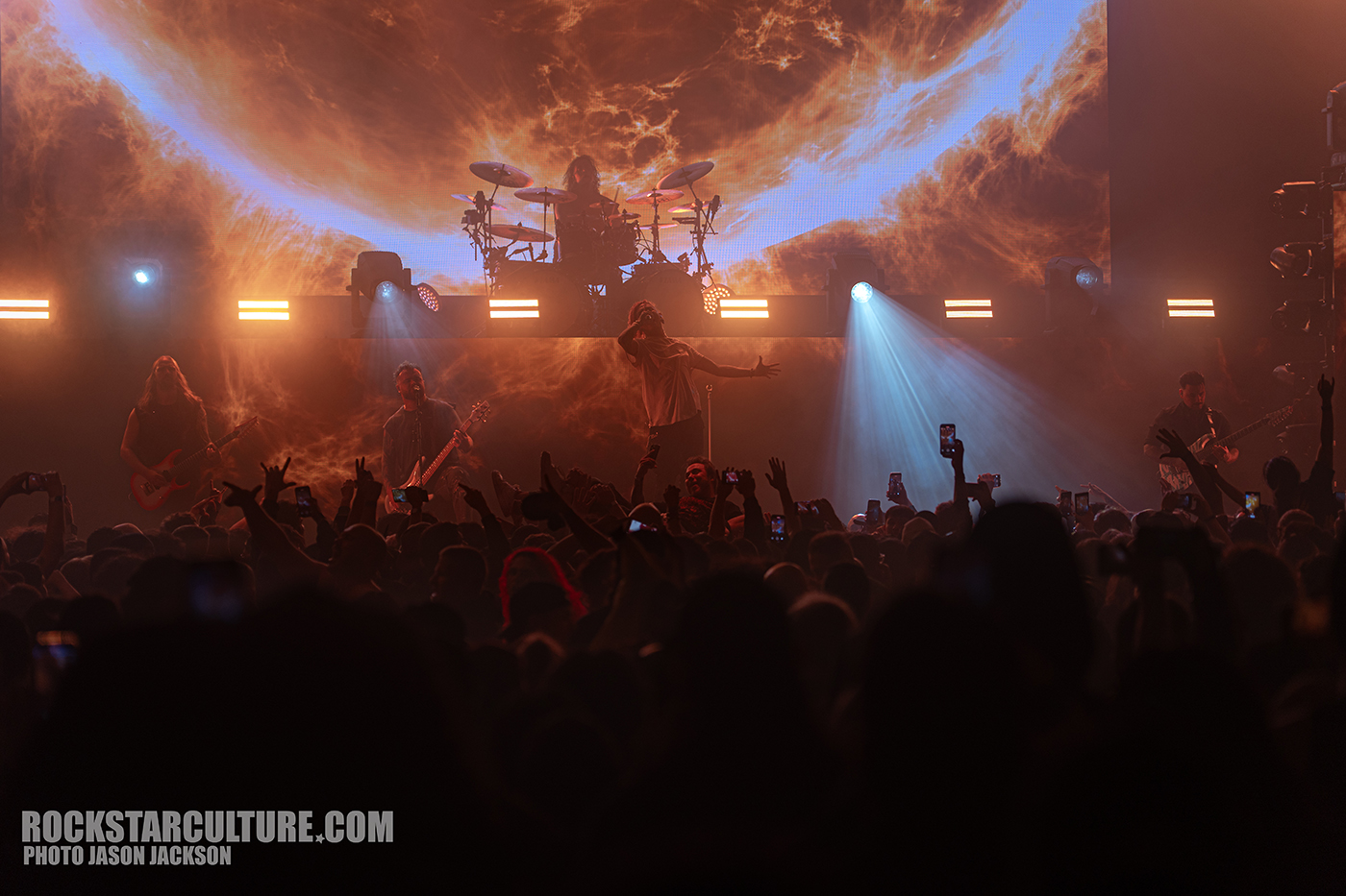 A lively concert scene with a band performing on stage in front of an audience with raised hands and phones. The backdrop features bright lighting and a dramatic design.