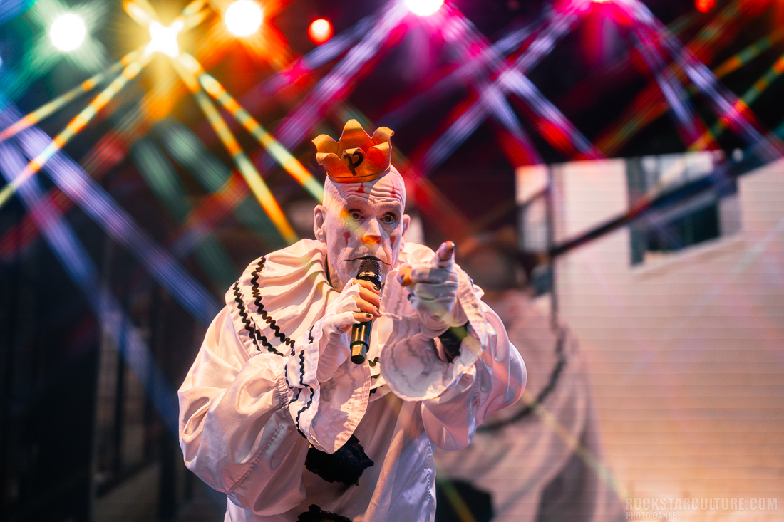 A performer dressed as a clown with white face paint and a crowned hat holds a microphone, creating an engaging atmosphere under colorful stage lights.