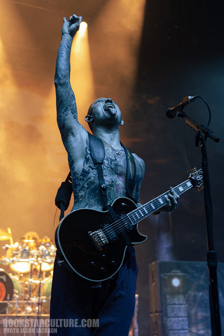 A tattooed musician passionately performing on stage, holding a guitar and raising one hand in the air, illuminated by dramatic stage lighting.