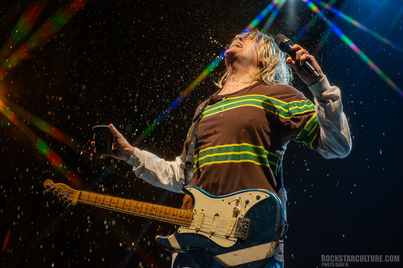 A musician on stage passionately performing with a guitar, water droplets sparkling around him, illuminated by colorful stage lights.