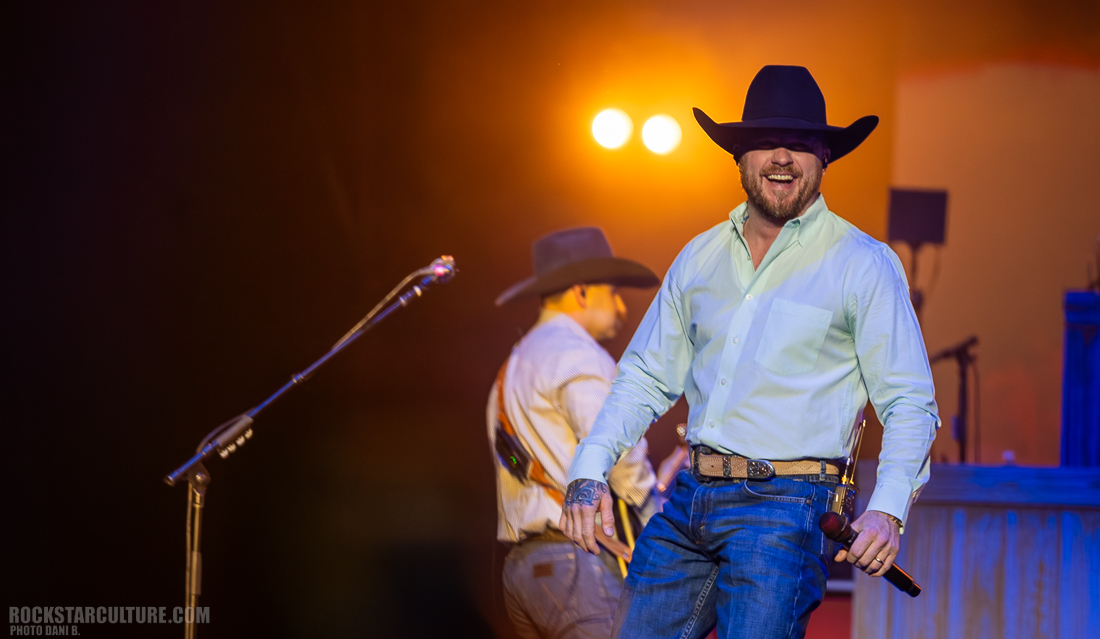 A smiling performer on stage wearing a cowboy hat and a light blue button-up shirt, holding a microphone. Another musician is partially visible in the background.