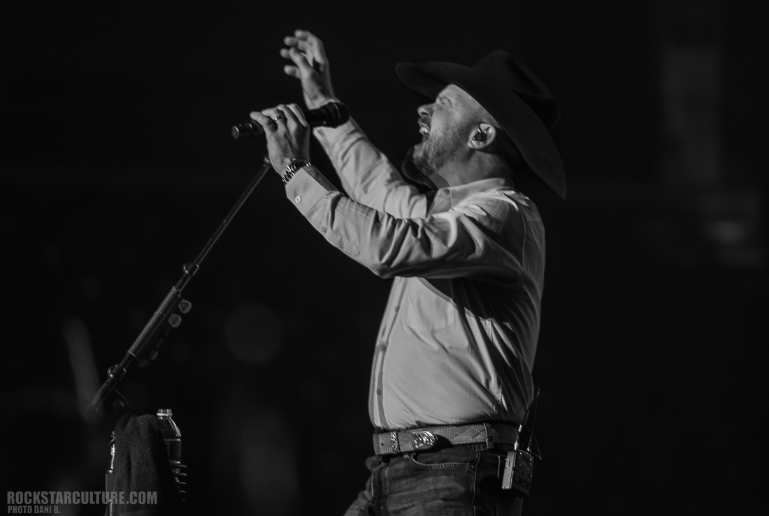 A singer passionately performing on stage, wearing a cowboy hat and holding a microphone, with dramatic lighting in a monochrome setting.
