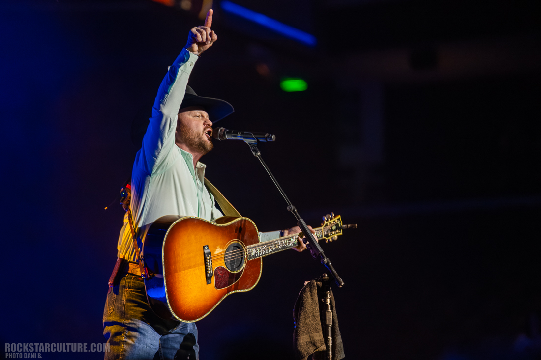 A singer performs on stage, holding a guitar and microphone, with one hand raised in the air, wearing a cowboy hat and a light blue button-up shirt.