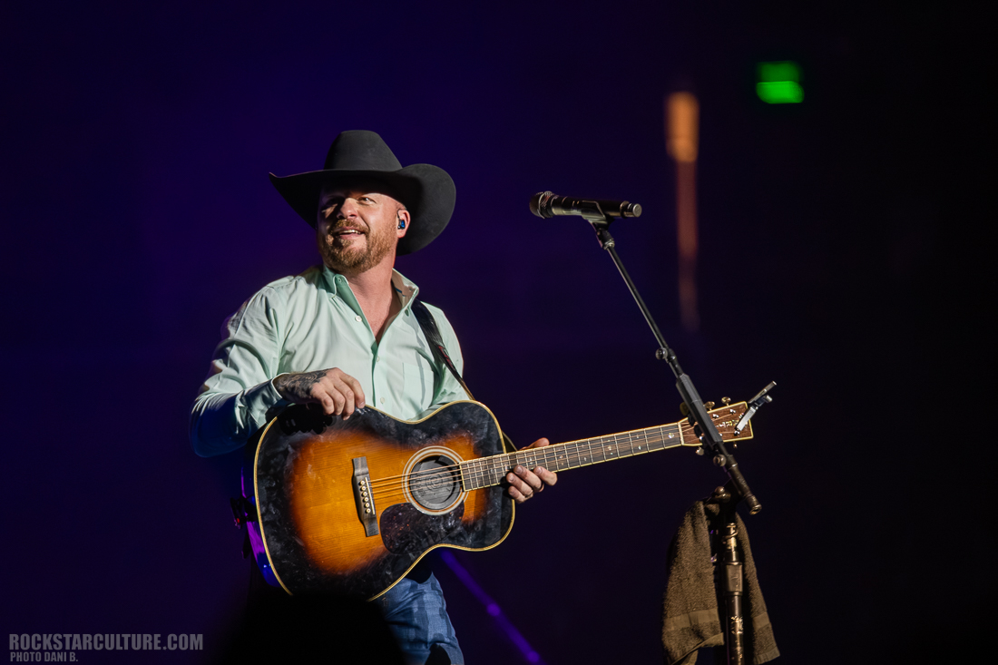 A country music artist wearing a hat and a light blue shirt is performing on stage with an acoustic guitar, smiling and engaging with the audience.