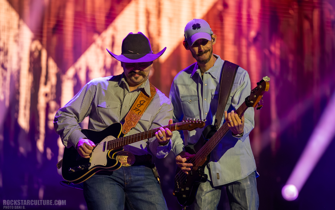 Two musicians performing on stage, one playing electric guitar and the other on bass, with a colorful background and bright stage lights.