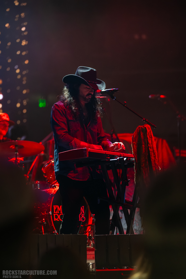 A musician playing a keyboard on stage, wearing a black hat and a red shirt, with a drummer visible in the background.