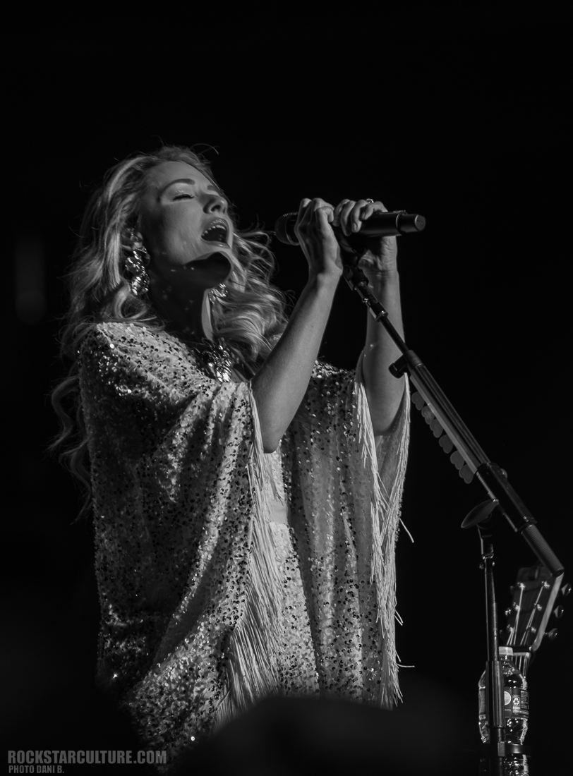 A female singer performing on stage, wearing a sparkling shawl and holding a microphone, captured in black and white.