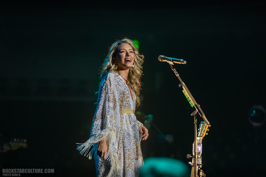 A female musician singing on stage wearing a white fringe outfit, with a microphone and guitar visible.