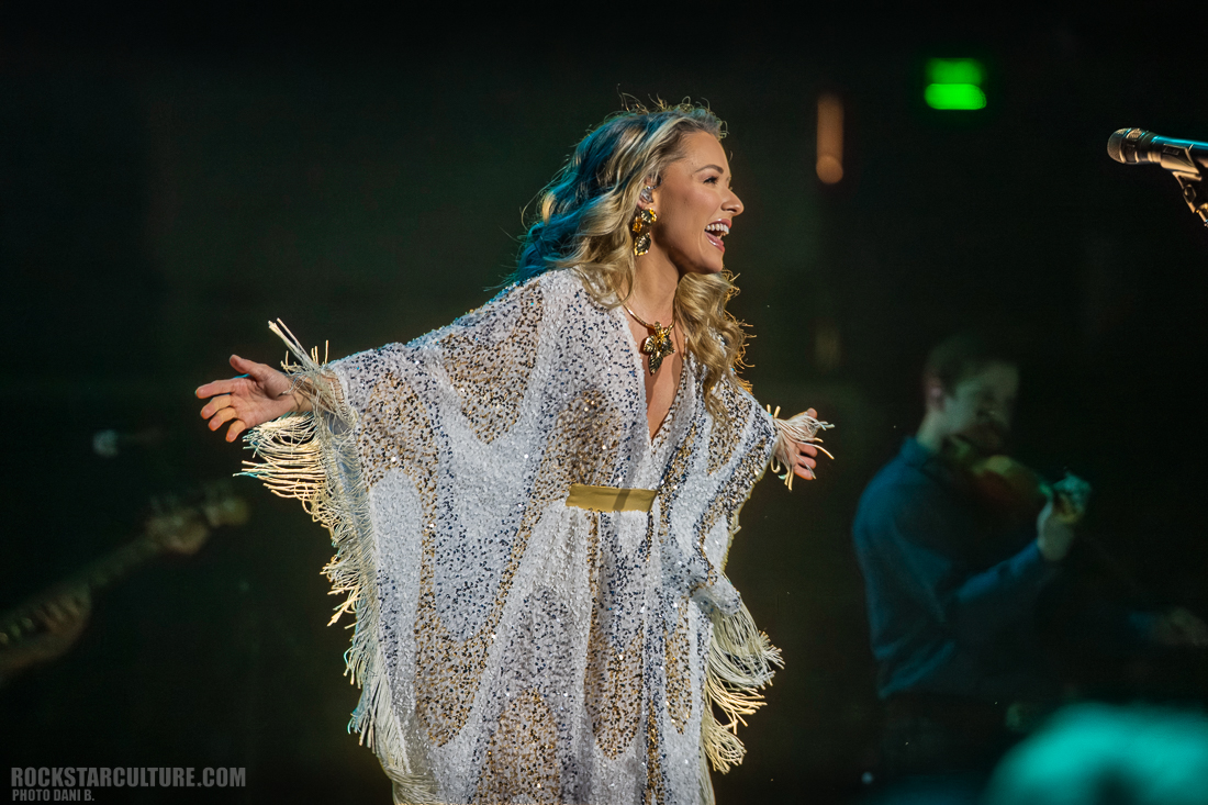 A female performer singing on stage, wearing a fringed white and gold outfit with a large necklace, smiling and gesturing with her hands. A musician playing a violin is slightly visible in the background.