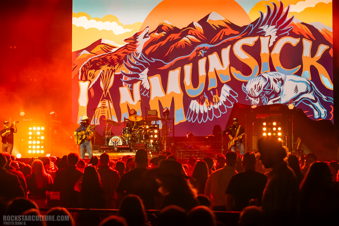 A country music band performing on stage in front of a large vibrant backdrop featuring the name 'Ian Munsick' with illustrations of nature, including mountains, an eagle, and a bison, alongside an audience enjoying the concert.