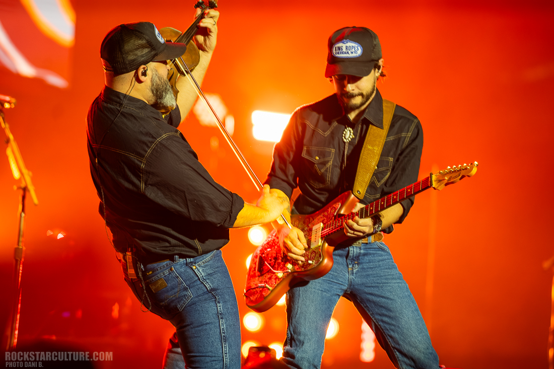 Two musicians performing on stage; one plays the violin while the other plays an electric guitar, both wearing black shirts and hats with illuminated background.