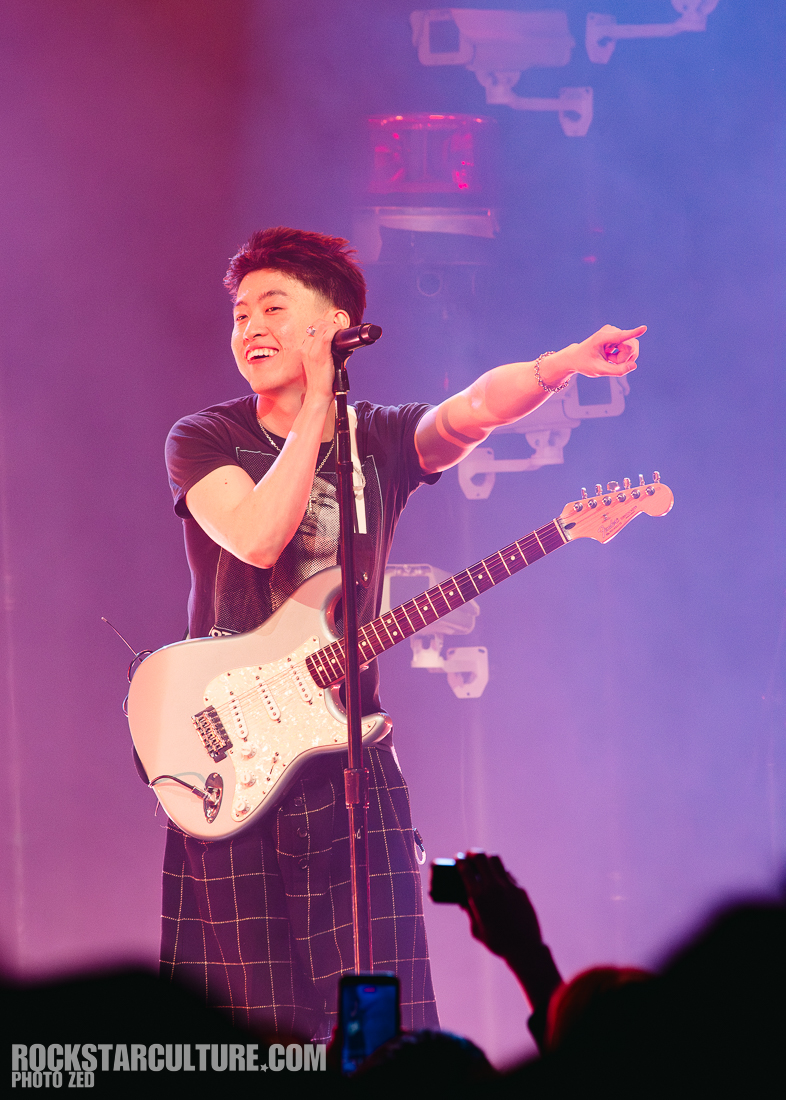 A musician smiles and points while performing on stage, holding an electric guitar. The background is filled with colorful stage lights and a lively atmosphere.