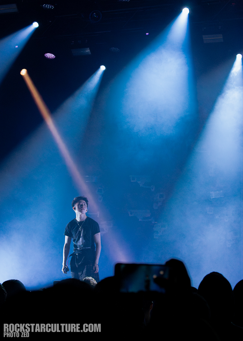 A performer stands on stage under dramatic lighting and smoke, engaging with the audience.