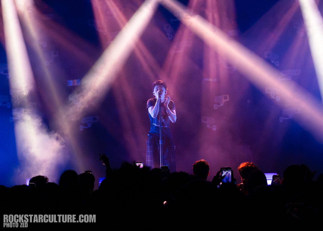 A singer performing on stage with a microphone, surrounded by dramatic lighting and a foggy atmosphere, while fans in the foreground watch and take photos.
