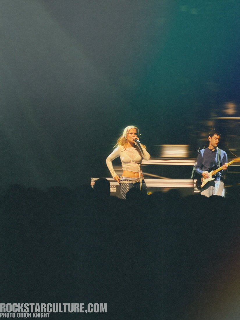 Singer performing on stage with a guitarist in the background, bright stage lights illuminating the scene.