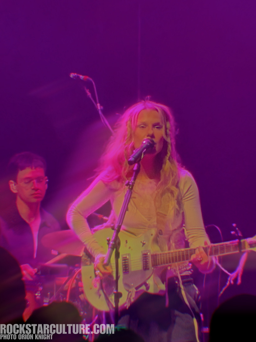 A female musician with long blonde hair playing an electric guitar and singing into a microphone on stage, with a drummer visible in the background, and colorful stage lighting creating a vibrant atmosphere.