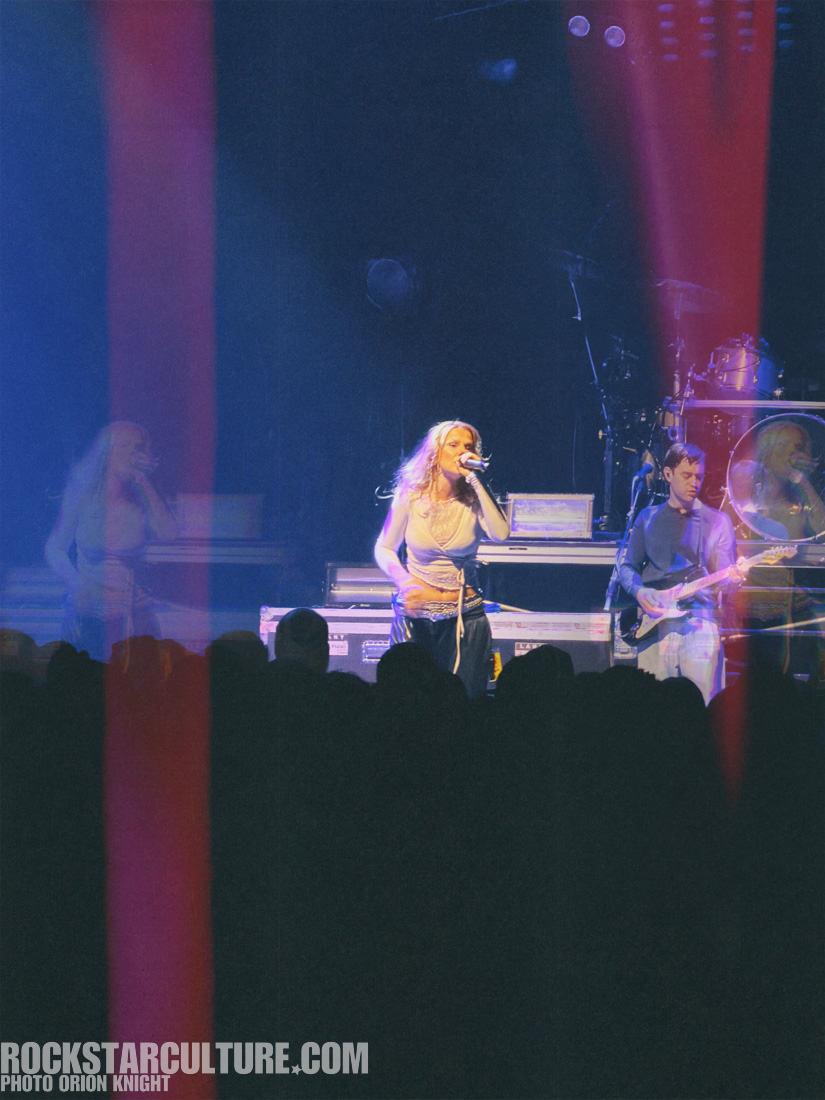 A singer performing on stage with a microphone, accompanied by a band, in a dimly lit concert setting with colorful stage lights.