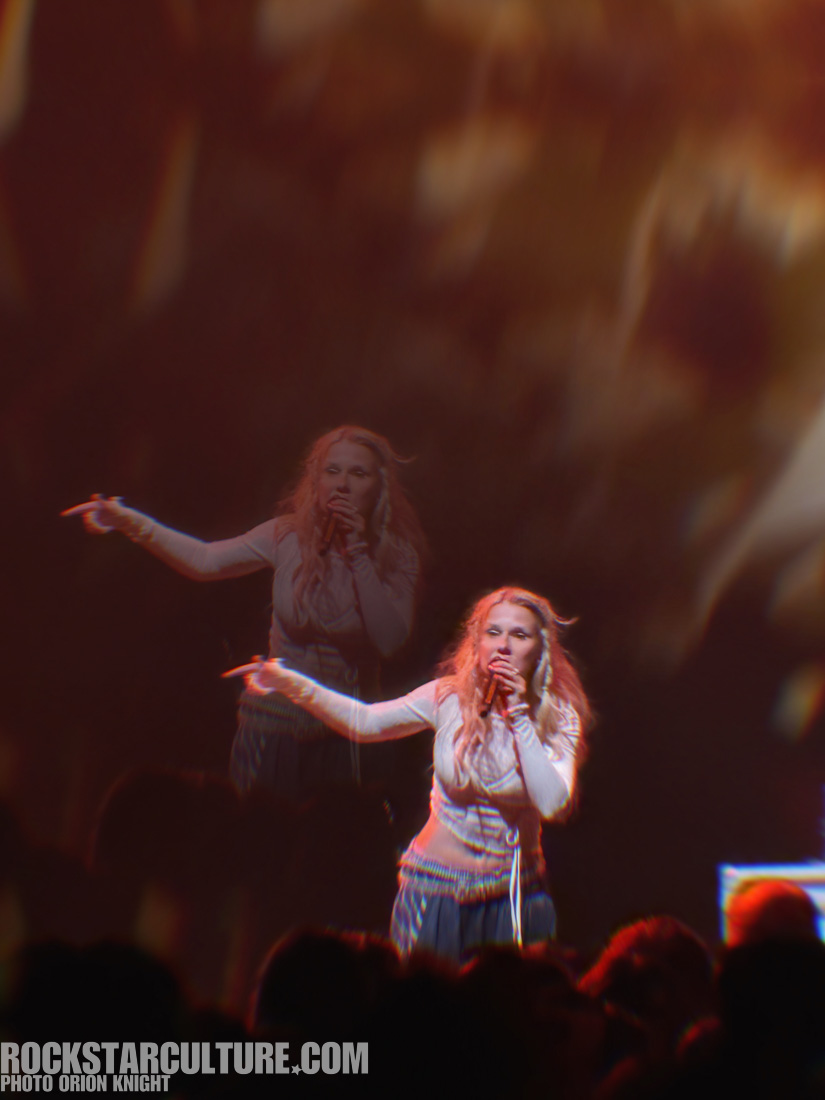 A female singer passionately performing on stage with a microphone, surrounded by a blurred, energetic crowd in a vibrant, colorful lighting atmosphere.