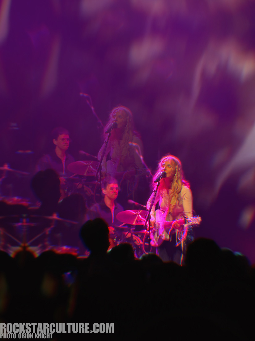 A female musician performing on stage with a microphone and guitar, accompanied by a male drummer, blurred crowd in the foreground and colorful stage lights in the background.