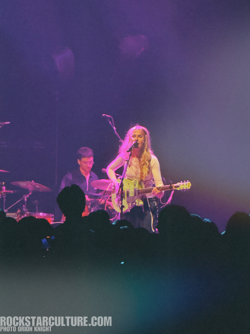 A female musician performing on stage with a guitar, while a drummer plays in the background, under colorful stage lighting.