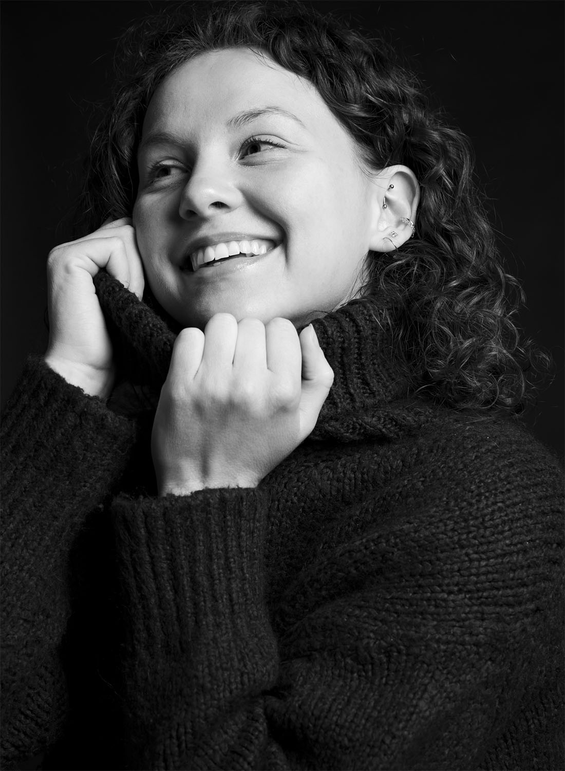 A smiling woman wearing a black sweater, holding the collar close to her face, with curly hair and minimal accessories, set against a dark background.