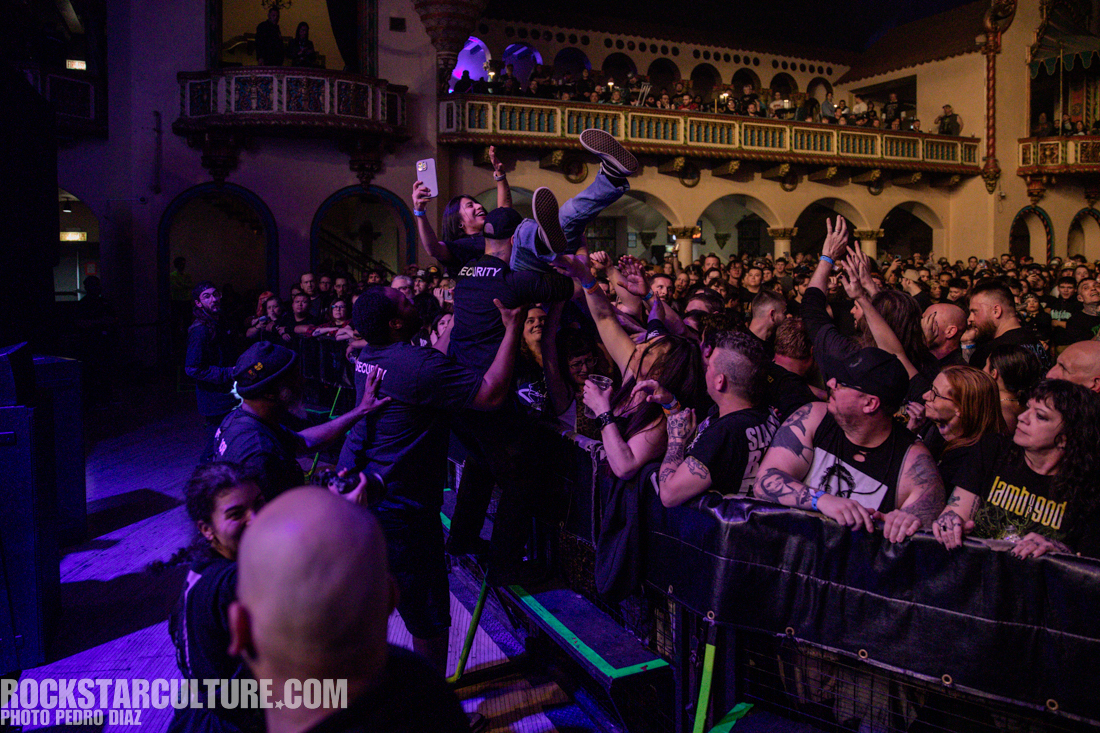 A lively concert scene with fans in the front row reaching out to catch a crowd surfer as security personnel assist. The audience is engaged and excited, with a variety of expressions visible.