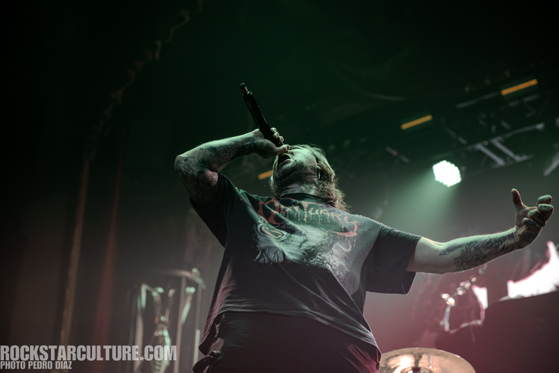 A male vocalist passionately performing on stage with a microphone, wearing a black graphic t-shirt, surrounded by a dramatic lighting backdrop.