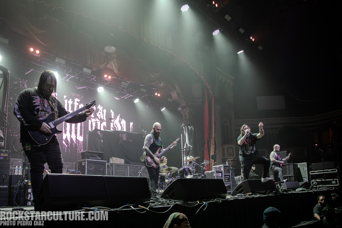 A live band performance with multiple musicians on stage, featuring guitars and vocals, set in a dimly lit venue with dramatic lighting.