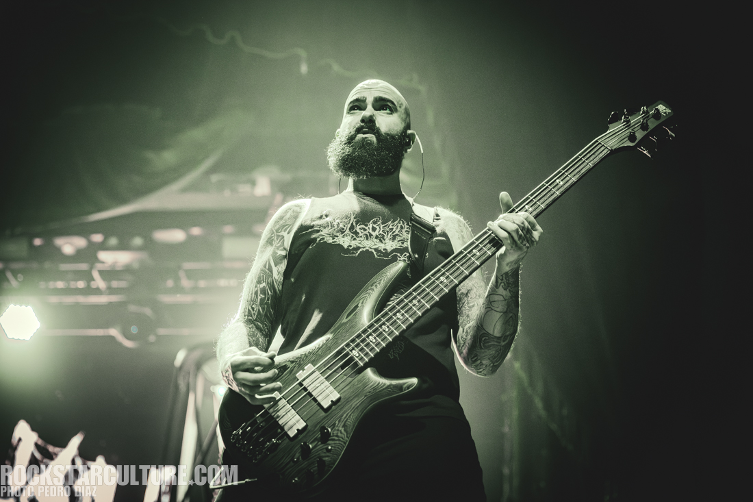 A musician with a beard playing a bass guitar on stage, dressed in a sleeveless black shirt with tattoos visible on their arms, under stage lighting.