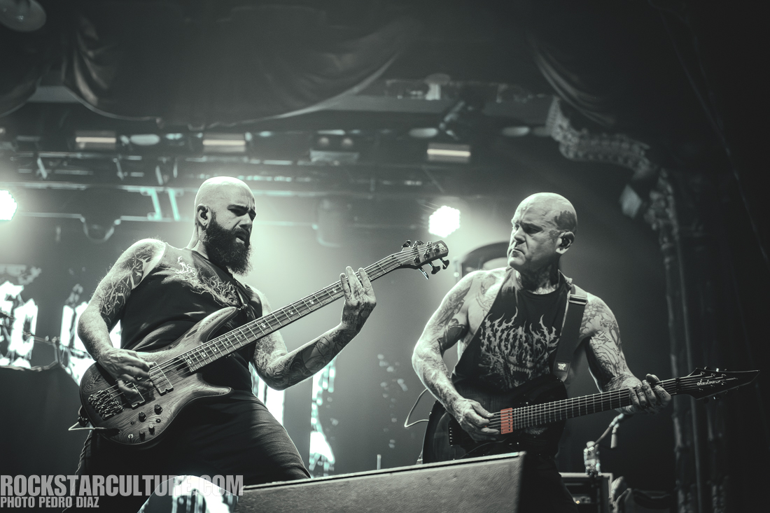 Two musicians performing on stage, one playing a bass guitar and the other playing an electric guitar, under dramatic stage lighting.