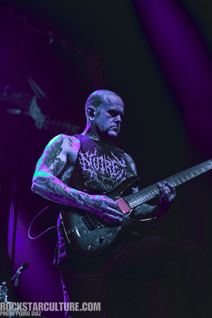 A tattooed guitarist playing an electric guitar on stage, under dramatic purple lighting.