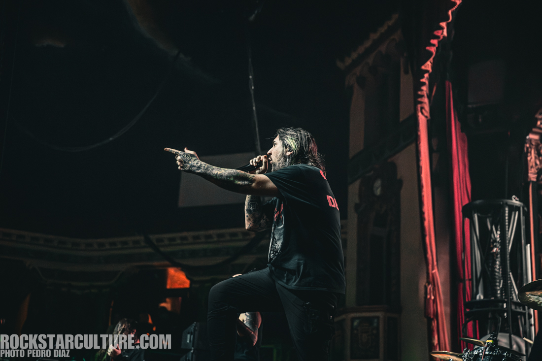 A male performer with tattoos passionately singing on stage, gesturing with one hand while holding a microphone in the other. The background features a dramatic stage setting with red curtains.