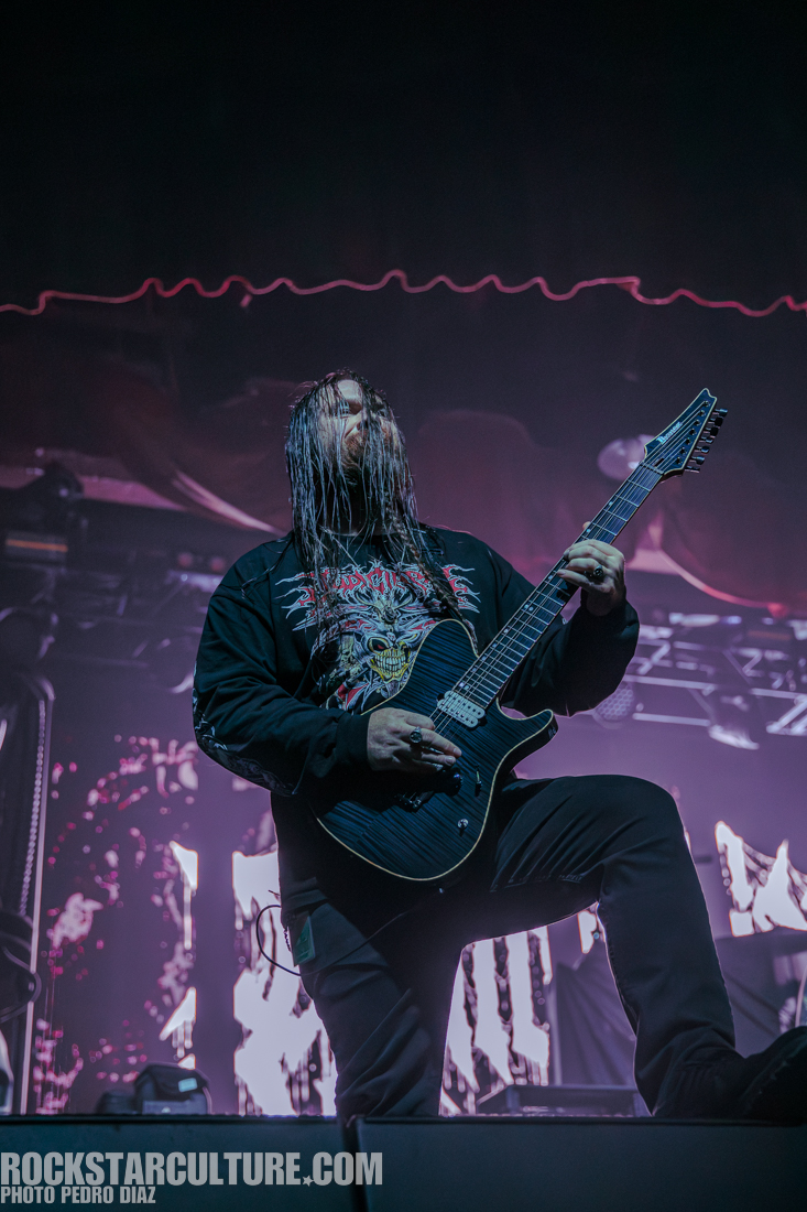 A musician performing on stage with long hair, wearing a graphic t-shirt and playing an electric guitar, with a dynamic lighting backdrop.