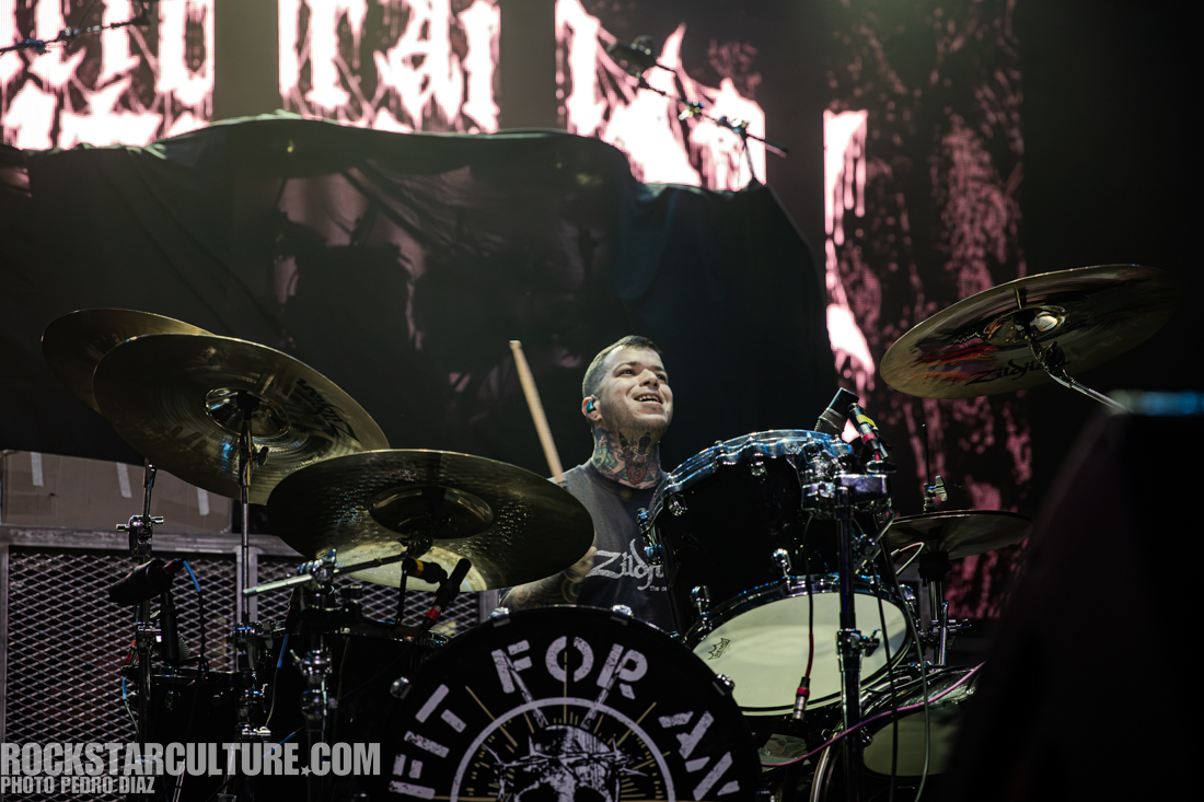 A drummer performing on stage, smiling and playing a drum set with various instruments and equipment in the background.
