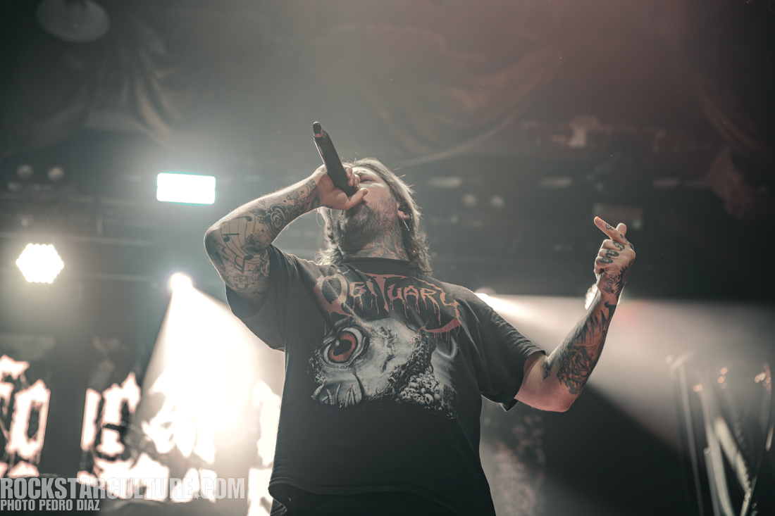 A man with tattoos performing on stage, wearing a black shirt with a graphic design, holding a microphone and gesturing towards the audience.