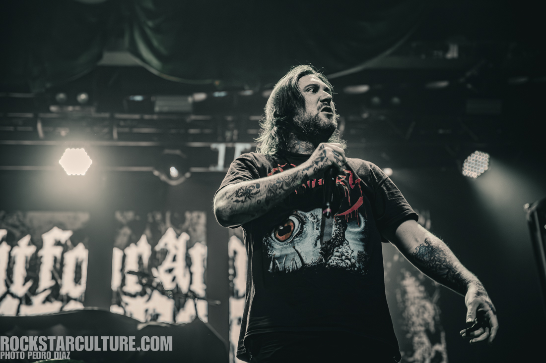 A male vocalist on stage wearing a black t-shirt with graphic designs. He is holding a microphone and has tattoos on his arms, with dramatic stage lighting in the background.