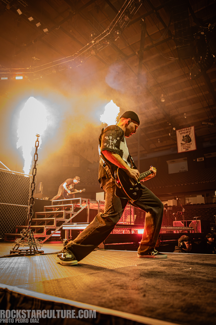 A guitarist performing on stage with dramatic lighting and pyrotechnics in the background, while another musician is visible in the distance.