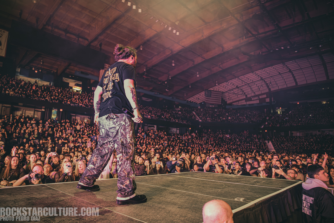 A performer stands on stage in front of a packed crowd at a concert, wearing a black shirt and camouflage pants. The audience, filled with fans taking photos, is illuminated by stage lights in a large indoor venue.