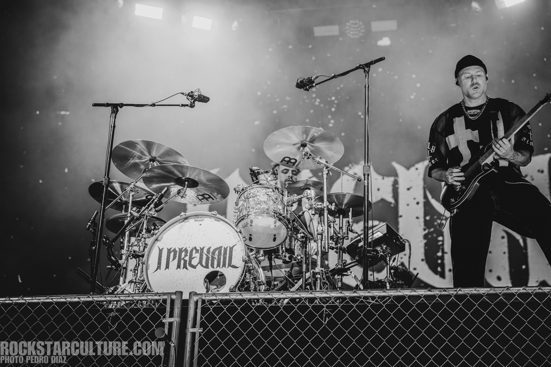 Black and white image of a live band performance featuring a drummer and a guitarist. The drummer is seated behind a drum kit with the band's name visible on the bass drum. The guitarist stands nearby, playing an electric guitar, with stage lights illuminating the background.