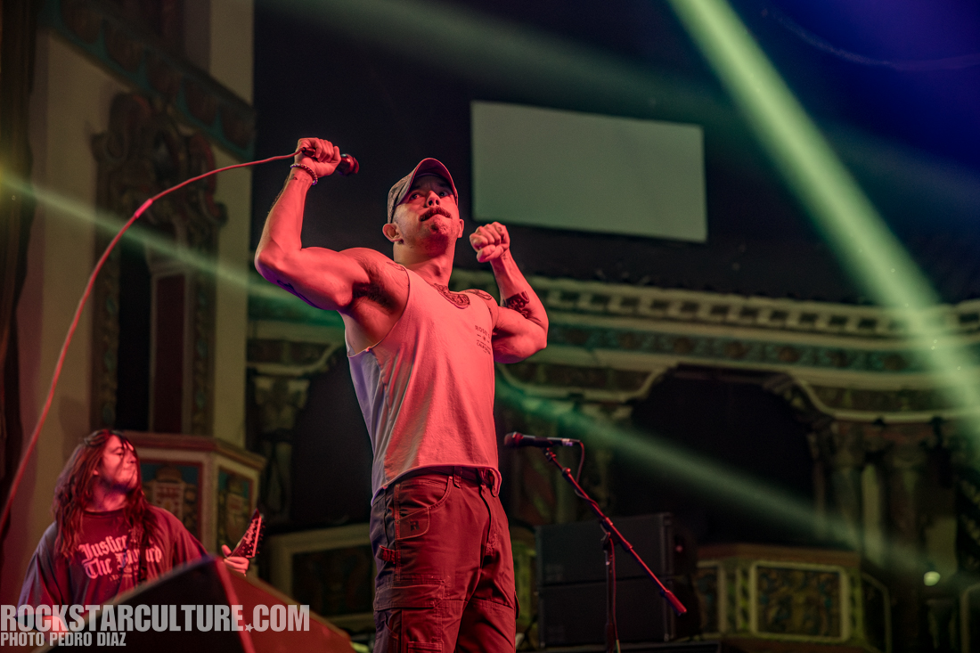 A male vocalist passionately performing on stage, muscular and in a sleeveless shirt, holding a microphone with vibrant stage lights in the background.