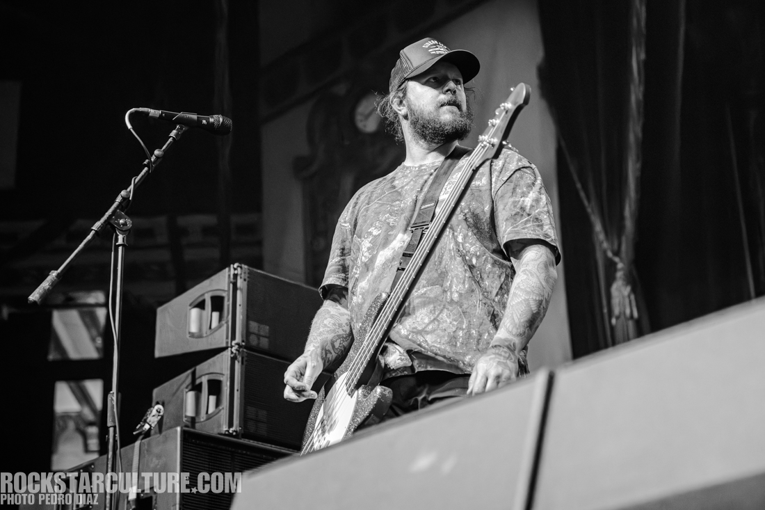 Musician performing on stage, playing a bass guitar while wearing a cap and a patterned shirt, with amplifiers in the background.