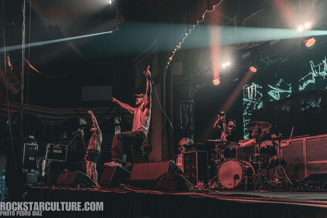 A live music performance featuring a band on stage, with the lead singer energetically engaging the audience. The drummer and another band member are also visible, showcasing a dynamic rock atmosphere with stage lights and equipment in the background.