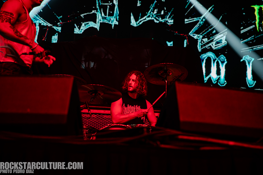 A drummer performing on stage with long curly hair, wearing a sleeveless black shirt, surrounded by a colorful backdrop. Another musician is partially visible in the foreground.