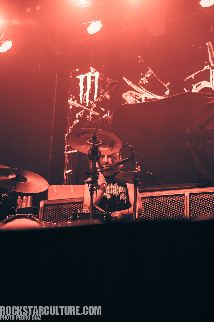 A drummer performing on stage with a dark background and red lighting, wearing a black tank top with a graphic design.
