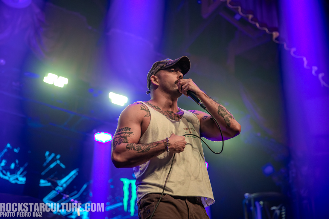 A male performer on stage wearing a sleeveless shirt and a cap, speaking into a microphone with vibrant stage lighting in the background.