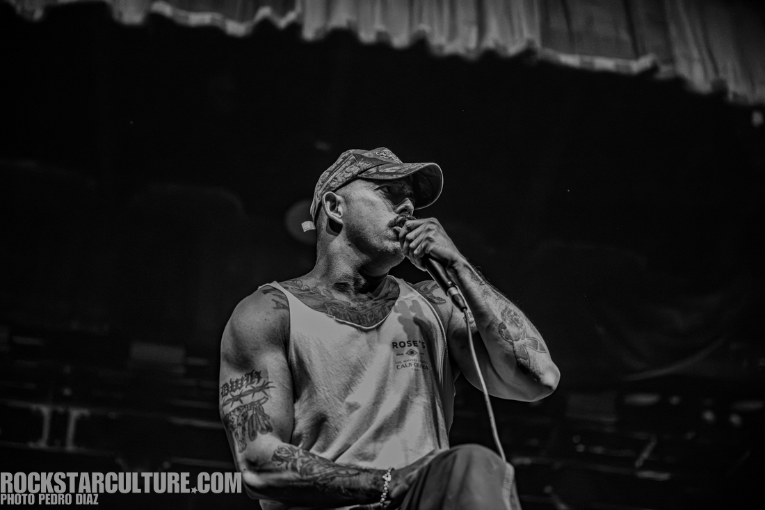 A muscular male performer singing into a microphone on stage, wearing a tank top and a cap, with visible tattoos on his arms.