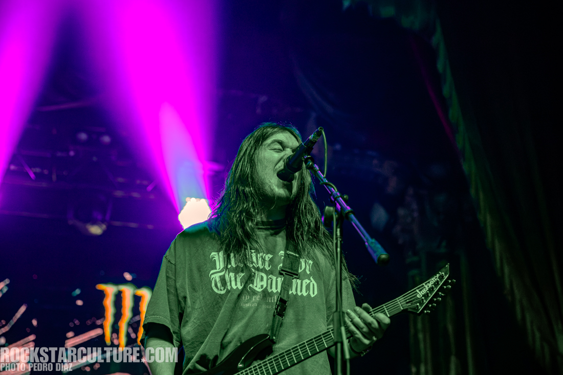A musician performing on stage with a guitar and microphone, wearing a graphic t-shirt. Colorful stage lights create an energetic atmosphere.
